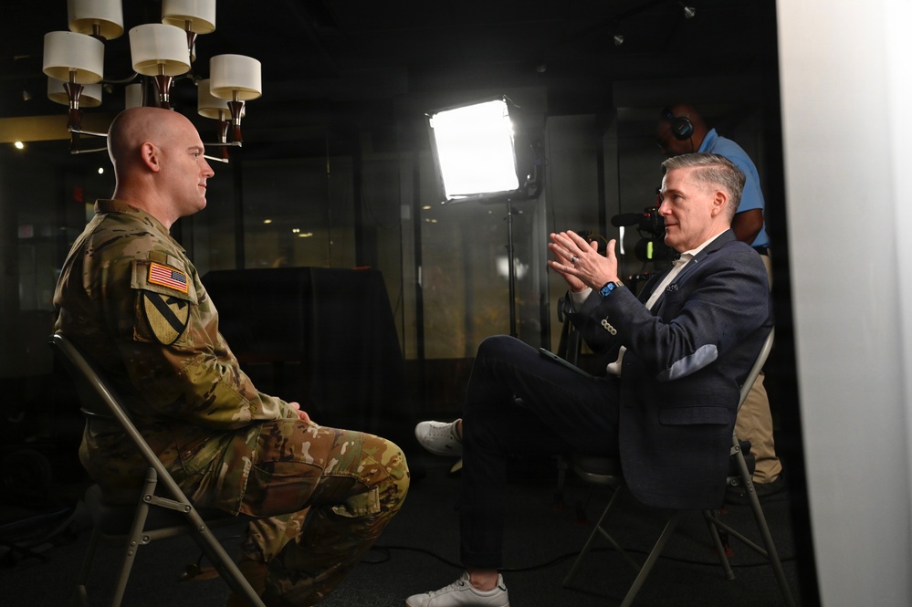 A U.S. Army soldier in camouflage uniform is being interviewed by a civilian man in a navy suit, seated facing each other in what appears to be a professional media setting. A bright LED panel light illuminates the subjects while a camera operator records the interview from behind. The setting suggests a formal media production or press interview environment.