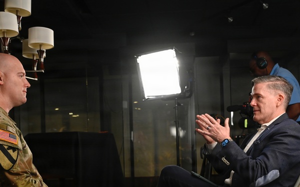 A U.S. Army soldier in camouflage uniform is being interviewed by a civilian man in a navy suit, seated facing each other in what appears to be a professional media setting. A bright LED panel light illuminates the subjects while a camera operator records the interview from behind. The setting suggests a formal media production or press interview environment.