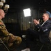 A U.S. Army soldier in camouflage uniform is being interviewed by a civilian man in a navy suit, seated facing each other in what appears to be a professional media setting. A bright LED panel light illuminates the subjects while a camera operator records the interview from behind. The setting suggests a formal media production or press interview environment.