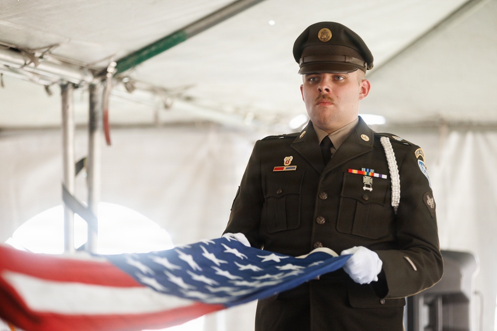 A U.S. Army soldier in dress uniform holds a partially folded American flag during what appears to be a military funeral or memorial ceremony. The soldier is wearing white gloves and stands at attention under a white tent, with a solemn and focused expression. The ceremony appears formal, consistent with military honors protocols.
