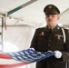 A U.S. Army soldier in dress uniform holds a partially folded American flag during what appears to be a military funeral or memorial ceremony. The soldier is wearing white gloves and stands at attention under a white tent, with a solemn and focused expression. The ceremony appears formal, consistent with military honors protocols.