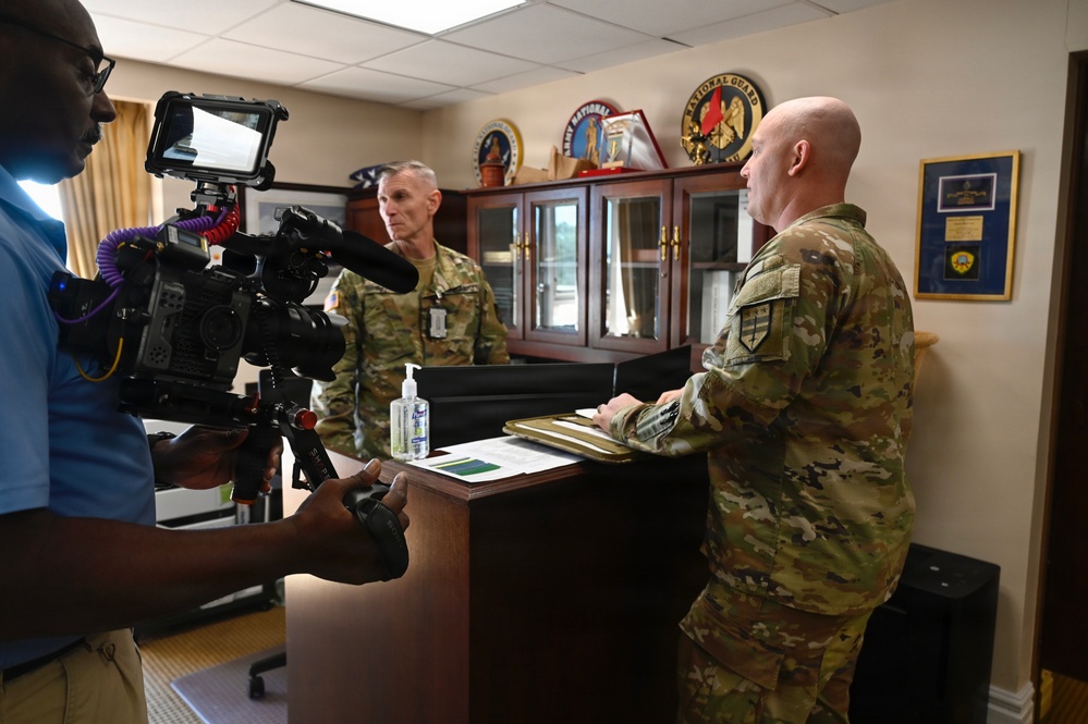 A videographer is filming two military personnel in camouflage uniforms inside what appears to be a military office. One soldier stands behind a desk holding a folder or clipboard, while another soldier stands across from him. The setting includes military insignia, plaques, and display cases visible in the background.