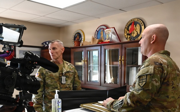 A videographer is filming two military personnel in camouflage uniforms inside what appears to be a military office. One soldier stands behind a desk holding a folder or clipboard, while another soldier stands across from him. The setting includes military insignia, plaques, and display cases visible in the background.