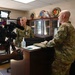 A videographer is filming two military personnel in camouflage uniforms inside what appears to be a military office. One soldier stands behind a desk holding a folder or clipboard, while another soldier stands across from him. The setting includes military insignia, plaques, and display cases visible in the background.