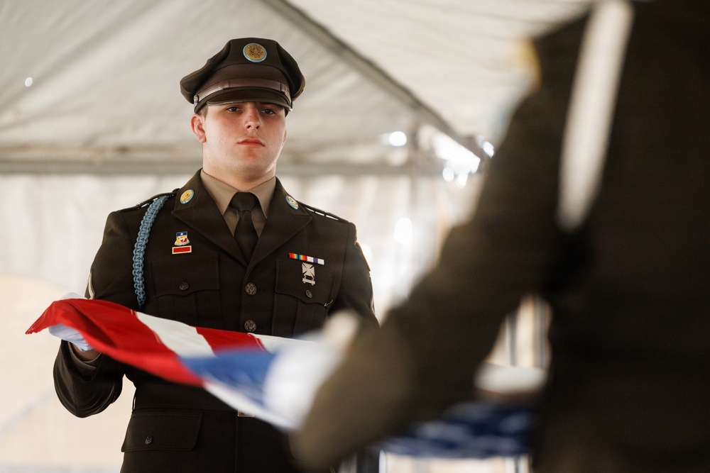 A young service member in a dark dress uniform is participating in a formal flag folding ceremony, likely a military or JROTC honor guard event. The individual holds a partially folded American flag with white-gloved hands while facing another participant across from them. The setting appears to be under a white tent or canopy, suggesting an outdoor ceremonial event.