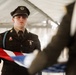 A young service member in a dark dress uniform is participating in a formal flag folding ceremony, likely a military or JROTC honor guard event. The individual holds a partially folded American flag with white-gloved hands while facing another participant across from them. The setting appears to be under a white tent or canopy, suggesting an outdoor ceremonial event.