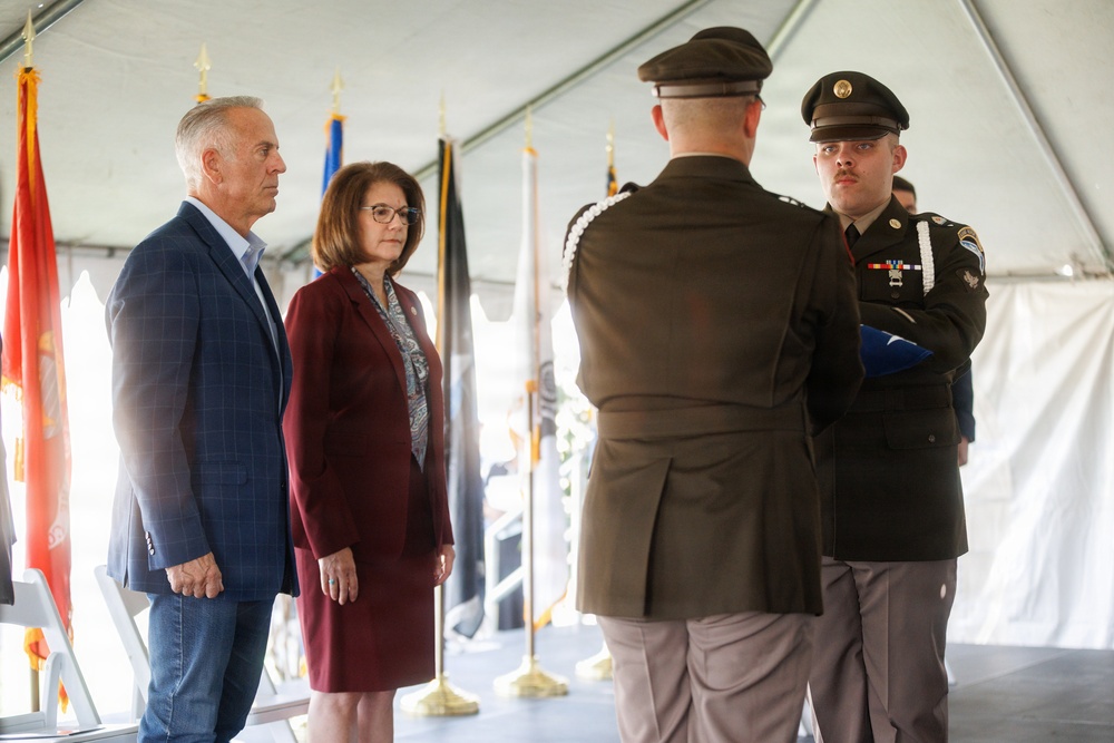 A formal military ceremony is taking place under a tent, where two uniformed soldiers are presenting a folded flag to two civilians — a man in a blue suit and a woman in a burgundy blazer. The setting includes multiple military branch flags in the background, suggesting an official government or memorial event. The solemn expressions and formal postures indicate this is likely a dignitary flag presentation or memorial service.