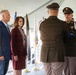 A formal military ceremony is taking place under a tent, where two uniformed soldiers are presenting a folded flag to two civilians — a man in a blue suit and a woman in a burgundy blazer. The setting includes multiple military branch flags in the background, suggesting an official government or memorial event. The solemn expressions and formal postures indicate this is likely a dignitary flag presentation or memorial service.