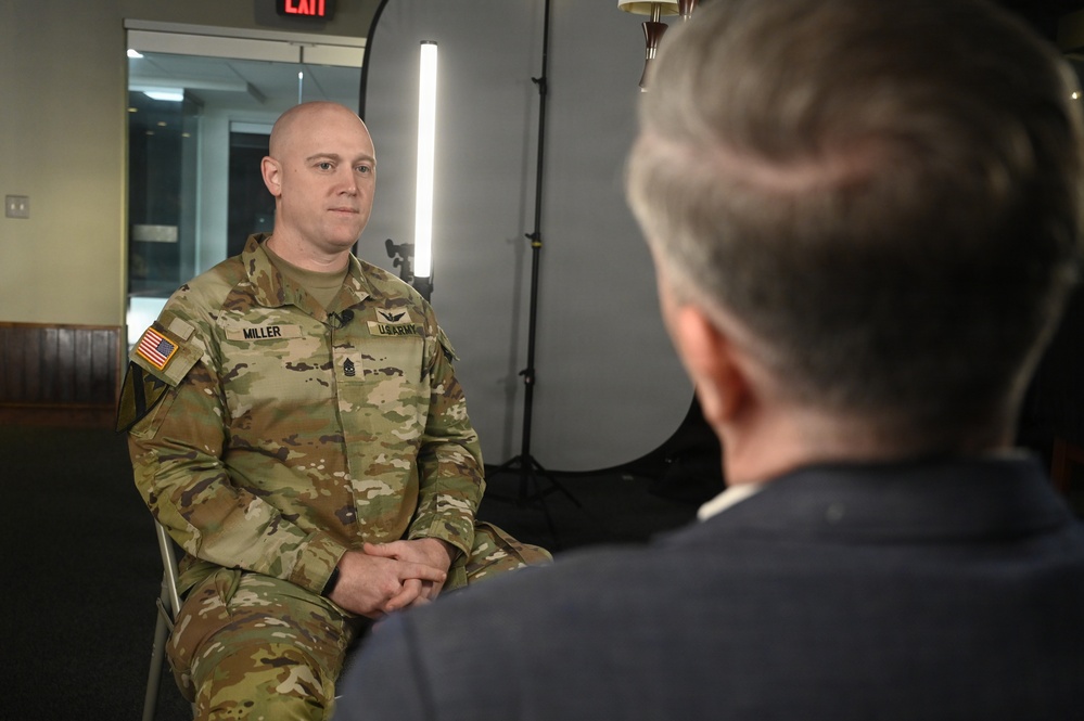 A U.S. Army soldier named Miller, wearing OCP (Operational Camouflage Pattern) uniform, is seated and participating in what appears to be a formal interview or media engagement. He is facing another individual whose back is to the camera, suggesting an interviewer-interviewee setup. The setting includes professional lighting equipment consistent with a media or press interview environment.