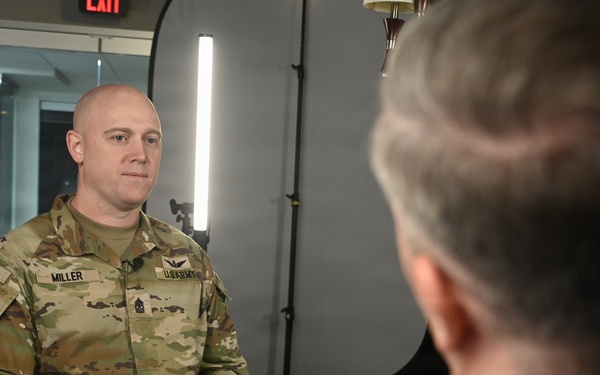 A U.S. Army soldier named Miller, wearing OCP (Operational Camouflage Pattern) uniform, is seated and participating in what appears to be a formal interview or media engagement. He is facing another individual whose back is to the camera, suggesting an interviewer-interviewee setup. The setting includes professional lighting equipment consistent with a media or press interview environment.