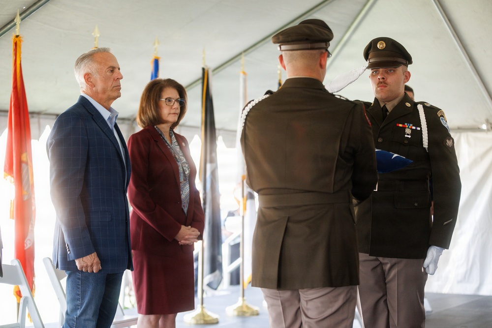 A military funeral or memorial ceremony is taking place under a white tent. Two uniformed soldiers, one holding a folded American flag, face two civilians — a man in a blue blazer and a woman in a burgundy suit — in what appears to be a flag presentation ceremony. Multiple ceremonial flags are visible in the background.