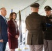 A military funeral or memorial ceremony is taking place under a white tent. Two uniformed soldiers, one holding a folded American flag, face two civilians — a man in a blue blazer and a woman in a burgundy suit — in what appears to be a flag presentation ceremony. Multiple ceremonial flags are visible in the background.