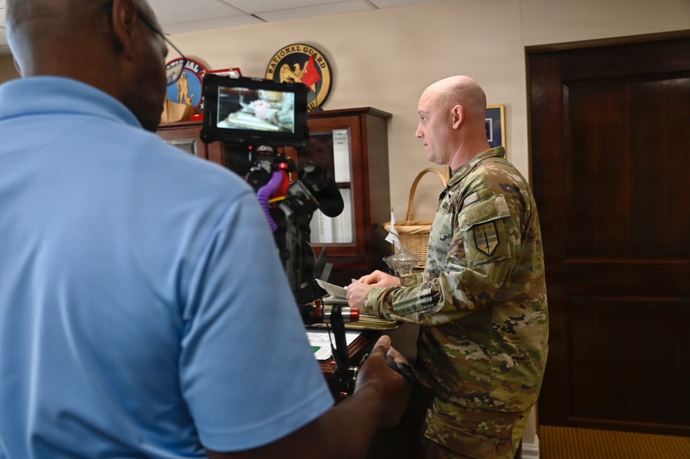 A military personnel in OCP (Operational Camouflage Pattern) uniform is being filmed or interviewed in what appears to be an office setting. A civilian cameraman in a blue polo shirt operates a camera rig with a monitor attached. The background displays military insignia including a visible National Guard emblem.