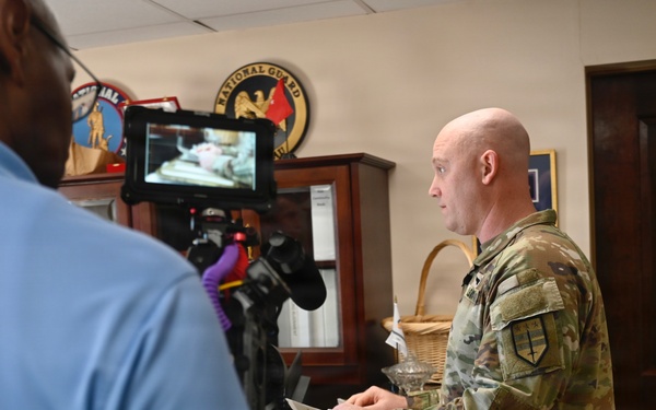 A military personnel in OCP (Operational Camouflage Pattern) uniform is being filmed or interviewed in what appears to be an office setting. A civilian cameraman in a blue polo shirt operates a camera rig with a monitor attached. The background displays military insignia including a visible National Guard emblem.