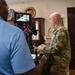 A military personnel in OCP (Operational Camouflage Pattern) uniform is being filmed or interviewed in what appears to be an office setting. A civilian cameraman in a blue polo shirt operates a camera rig with a monitor attached. The background displays military insignia including a visible National Guard emblem.