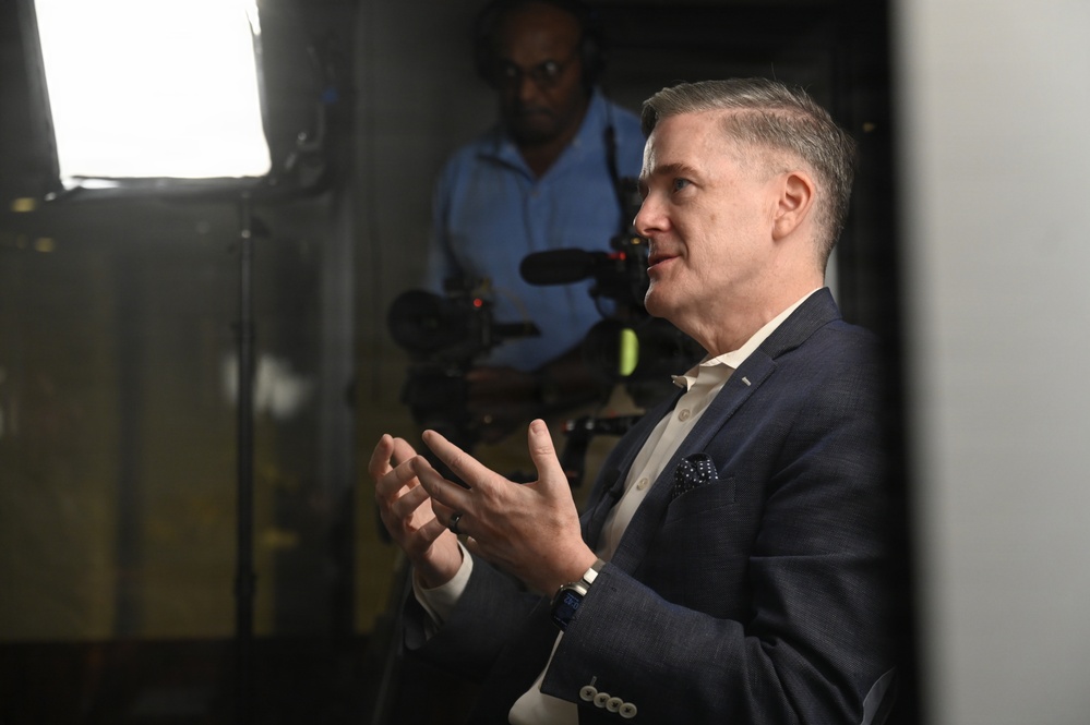 A middle-aged man in a navy blue blazer and white shirt is speaking or being interviewed, gesturing with both hands in an expressive manner. Behind him, a videographer in a blue shirt operates a professional camera with a mounted microphone, illuminated by a bright studio light panel. The setting appears to be a professional media or press environment.