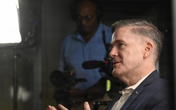 A middle-aged man in a navy blue blazer and white shirt is speaking or being interviewed, gesturing with both hands in an expressive manner. Behind him, a videographer in a blue shirt operates a professional camera with a mounted microphone, illuminated by a bright studio light panel. The setting appears to be a professional media or press environment.