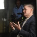 A middle-aged man in a navy blue blazer and white shirt is speaking or being interviewed, gesturing with both hands in an expressive manner. Behind him, a videographer in a blue shirt operates a professional camera with a mounted microphone, illuminated by a bright studio light panel. The setting appears to be a professional media or press environment.