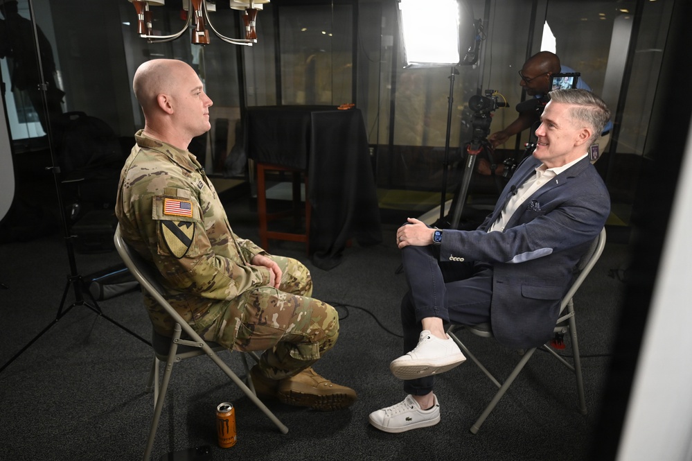 A U.S. Army soldier in OCP (Operational Camouflage Pattern) uniform is being interviewed by a civilian man in a navy blazer, both seated on folding chairs facing each other. A camera operator is visible in the background operating a professional video camera under studio lighting. The setting appears to be a professional media or broadcast interview setup.