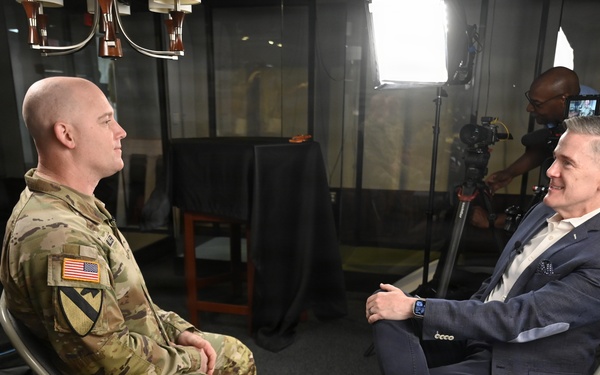 A U.S. Army soldier in OCP (Operational Camouflage Pattern) uniform is being interviewed by a civilian man in a navy blazer, both seated on folding chairs facing each other. A camera operator is visible in the background operating a professional video camera under studio lighting. The setting appears to be a professional media or broadcast interview setup.