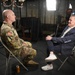 A U.S. Army soldier in OCP (Operational Camouflage Pattern) uniform is being interviewed by a civilian man in a navy blazer, both seated on folding chairs facing each other. A camera operator is visible in the background operating a professional video camera under studio lighting. The setting appears to be a professional media or broadcast interview setup.