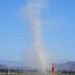 A dust devil — a small, rotating column of dust and debris — rises vertically from an arid, flat terrain in what appears to be a desert or semi-arid region. The scene includes colorful event flags in the foreground, suggesting a nearby outdoor event or venue. A mountain range is visible in the background under a clear blue sky.