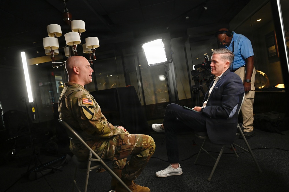 A U.S. Army soldier in OCP (Operational Camouflage Pattern) uniform is being interviewed by a civilian in a navy blazer in what appears to be a professional media setting. A camera operator in a blue polo shirt adjusts equipment in the background. The scene is lit with professional studio lighting equipment including a large LED panel and a vertical light strip.