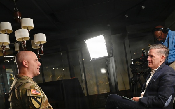 A U.S. Army soldier in OCP (Operational Camouflage Pattern) uniform is being interviewed by a civilian in a navy blazer in what appears to be a professional media setting. A camera operator in a blue polo shirt adjusts equipment in the background. The scene is lit with professional studio lighting equipment including a large LED panel and a vertical light strip.