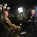 A U.S. Army soldier in OCP (Operational Camouflage Pattern) uniform is being interviewed by a civilian in a navy blazer in what appears to be a professional media setting. A camera operator in a blue polo shirt adjusts equipment in the background. The scene is lit with professional studio lighting equipment including a large LED panel and a vertical light strip.