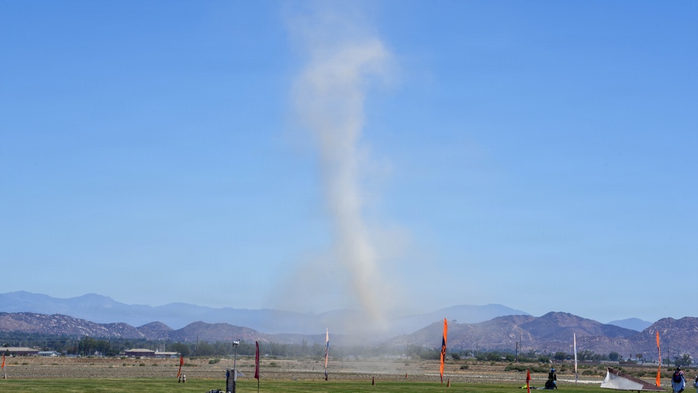 A dust devil or small whirlwind is visible rising from an open field at what appears to be an airfield or recreational flying area. The column of dust and debris extends high into a clear blue sky, with mountain ranges visible in the background. Orange marker flags and windsocks are positioned across the field, suggesting an organized aviation or kite-flying event.