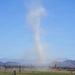 A dust devil or small whirlwind is visible rising from an open field at what appears to be an airfield or recreational flying area. The column of dust and debris extends high into a clear blue sky, with mountain ranges visible in the background. Orange marker flags and windsocks are positioned across the field, suggesting an organized aviation or kite-flying event.