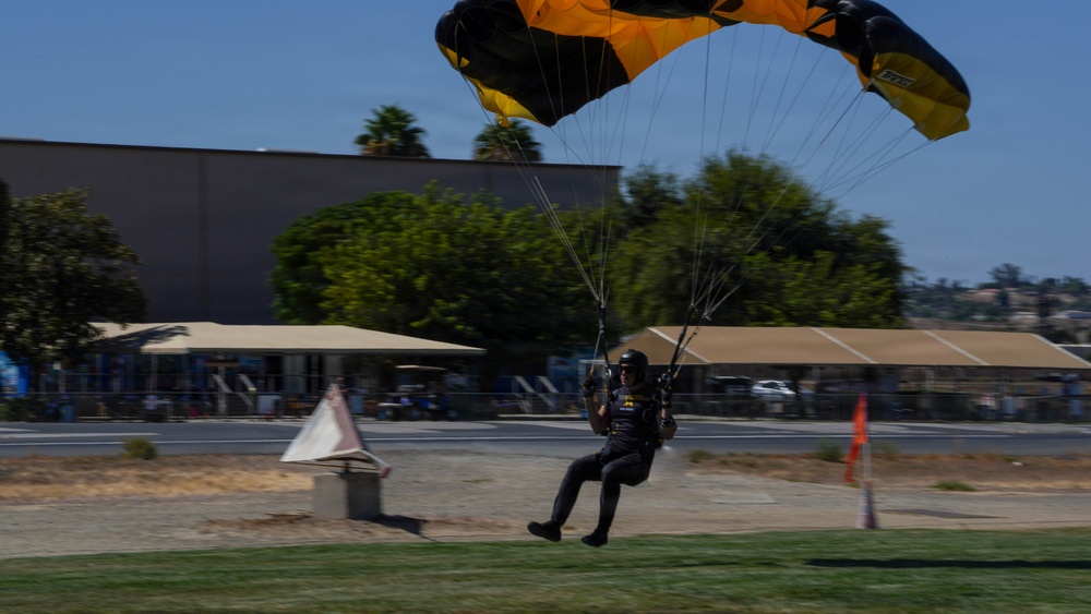 A skydiver is captured moments before landing on a grassy area, deploying a black and yellow parachute canopy. The setting appears to be a public airshow or demonstration event, with spectator tents, buildings, and trees visible in the background. The image has slight motion blur, suggesting the skydiver is moving at speed during the final approach.