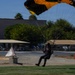 A skydiver is captured moments before landing on a grassy area, deploying a black and yellow parachute canopy. The setting appears to be a public airshow or demonstration event, with spectator tents, buildings, and trees visible in the background. The image has slight motion blur, suggesting the skydiver is moving at speed during the final approach.