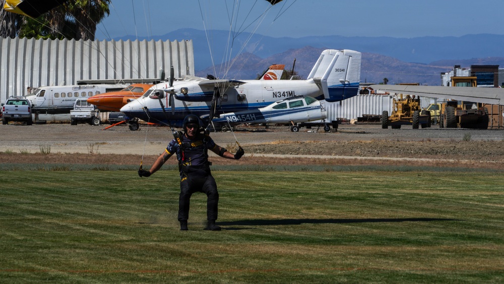 A skydiver is landing on a grass area at a small airfield or drop zone, with parachute lines visible above. Several aircraft are parked in the background near a hangar, along with ground vehicles and heavy equipment. The setting appears to be a rural or semi-arid airfield with mountains visible in the distance.
