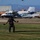 Canopy Relative Work Teams Compete at 2026 National Championships A skydiver is landing on a grass area at a small airfield or drop zone, with parachute lines visible above. Several aircraft are parked in the background near a hangar, along with ground vehicles and heavy equipment. The setting appears to be a rural or semi-arid airfield with mountains visible in the distance.