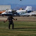 A skydiver is landing on a grass area at a small airfield or drop zone, with parachute lines visible above. Several aircraft are parked in the background near a hangar, along with ground vehicles and heavy equipment. The setting appears to be a rural or semi-arid airfield with mountains visible in the distance.