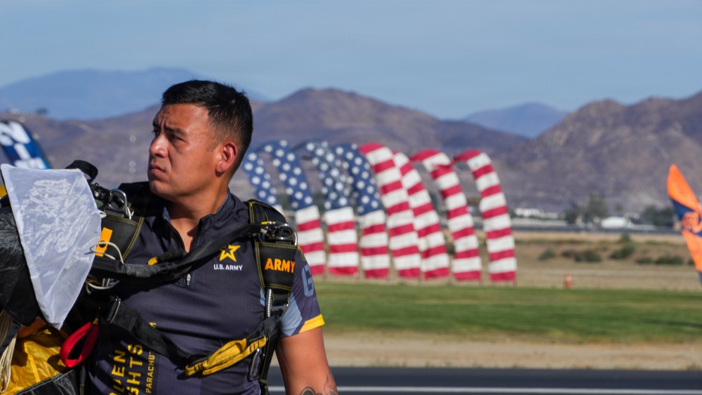 A U.S. Army parachutist, identifiable by his 'Golden Knights Parachute' team uniform, stands on an airfield tarmac carrying parachute equipment. In the background, multiple large American flag-shaped inflatables or sculptures are displayed in a row, set against a mountainous desert landscape. The scene appears to be at an airshow or military demonstration event.