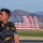 Canopy Relative Work Teams Compete at 2026 National Championships A U.S. Army parachutist, identifiable by his 'Golden Knights Parachute' team uniform, stands on an airfield tarmac carrying parachute equipment. In the background, multiple large American flag-shaped inflatables or sculptures are displayed in a row, set against a mountainous desert landscape. The scene appears to be at an airshow or military demonstration event.