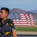 A U.S. Army parachutist, identifiable by his 'Golden Knights Parachute' team uniform, stands on an airfield tarmac carrying parachute equipment. In the background, multiple large American flag-shaped inflatables or sculptures are displayed in a row, set against a mountainous desert landscape. The scene appears to be at an airshow or military demonstration event.