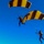 Golden Knights Defend Canopy Formation Title at Perris Two skydivers are descending under fully deployed parachutes against a clear blue sky. Both canopies share an identical yellow and dark brown/black striped pattern, suggesting they are part of the same team or demonstration group. The image is taken from below and slightly to the side, capturing both jumpers in close proximity during their descent.