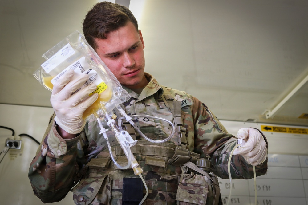 A U.S. Army soldier wearing OCP (Operational Camouflage Pattern) uniform and latex gloves is handling an IV fluid bag with attached tubing and connectors. The soldier appears to be preparing or inspecting intravenous medical equipment, likely in a field medical or military treatment facility setting. A yellow label is visible on the IV bag, and the soldier is concentrating on the tubing connections.