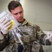 A U.S. Army soldier wearing OCP (Operational Camouflage Pattern) uniform and latex gloves is handling an IV fluid bag with attached tubing and connectors. The soldier appears to be preparing or inspecting intravenous medical equipment, likely in a field medical or military treatment facility setting. A yellow label is visible on the IV bag, and the soldier is concentrating on the tubing connections.