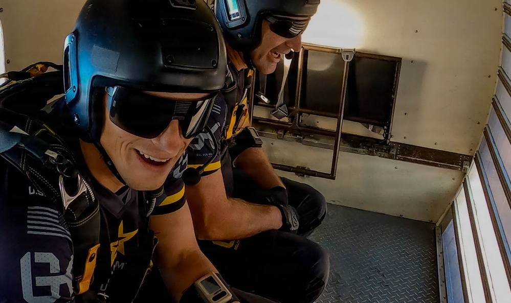 Two individuals in full skydiving gear are seated inside what appears to be an aircraft, preparing for a jump. Both are wearing helmets, goggles, jumpsuits with black and yellow markings, and parachute rigs. They appear to be smiling and in good spirits, suggesting anticipation before a jump.