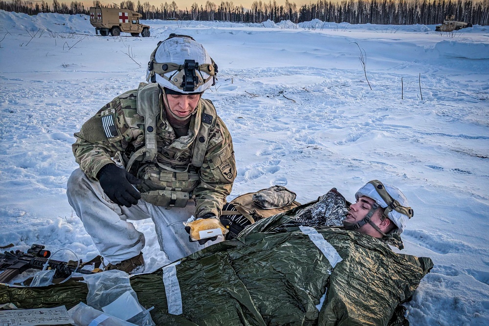 A U.S. Army soldier in multicam/ACU winter combat gear administers field medical treatment to a simulated casualty lying on the snow-covered ground wrapped in a thermal emergency blanket. The scene appears to be a cold-weather combat casualty care training exercise, with military medical vehicles visible in the background. The setting is a snow-covered open area bordered by a treeline, consistent with an Alaskan or northern training environment.
