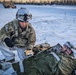A U.S. Army soldier in multicam/ACU winter combat gear administers field medical treatment to a simulated casualty lying on the snow-covered ground wrapped in a thermal emergency blanket. The scene appears to be a cold-weather combat casualty care training exercise, with military medical vehicles visible in the background. The setting is a snow-covered open area bordered by a treeline, consistent with an Alaskan or northern training environment.