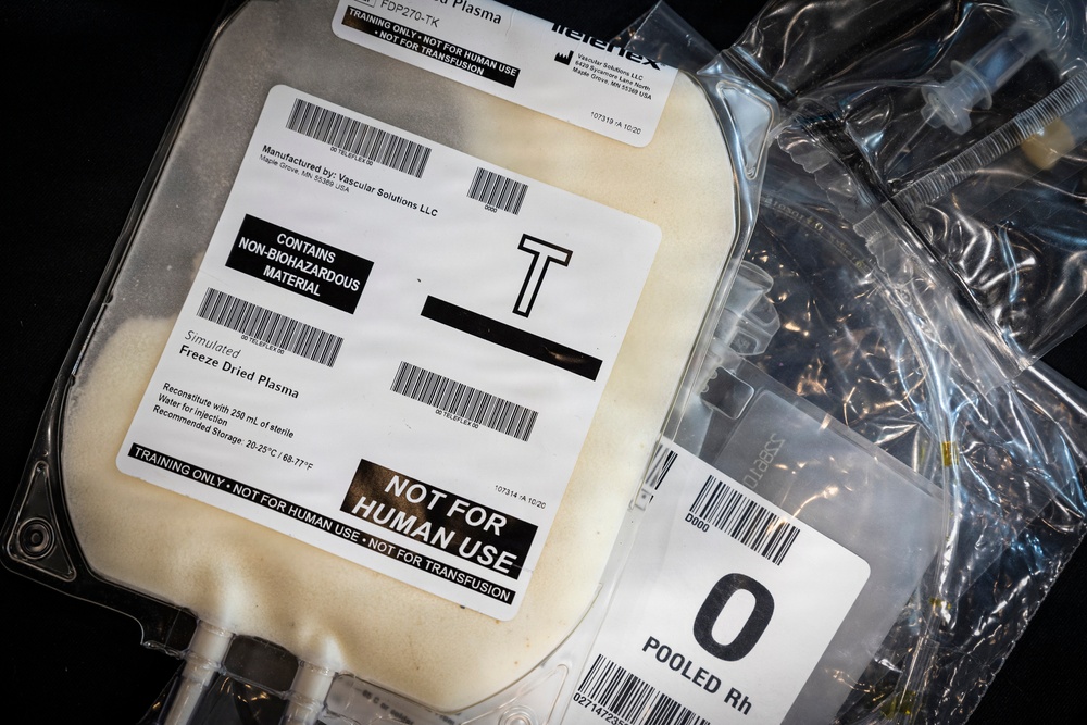 The image shows medical training supplies, specifically simulated freeze-dried plasma bags used for training purposes only. The primary bag is labeled with multiple warnings indicating it is 'NOT FOR HUMAN USE' and 'NOT FOR TRANSFUSION,' and contains non-biohazardous material. A secondary bag visible on the right is labeled 'O POOLED Rh,' suggesting it simulates Type O Rh blood product for training scenarios.