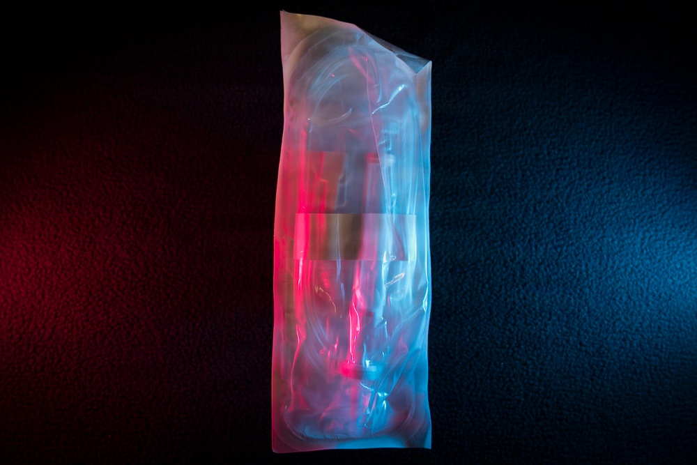 A transparent plastic bag or packaging is photographed against a dark textured background, illuminated by contrasting red and blue colored lighting. The dual-tone lighting creates a dramatic visual effect, with the left side bathed in red/pink light and the right side in cyan/blue light. The bag appears to contain some coiled or bundled items, though the exact contents are difficult to discern through the translucent material.