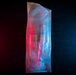 A transparent plastic bag or packaging is photographed against a dark textured background, illuminated by contrasting red and blue colored lighting. The dual-tone lighting creates a dramatic visual effect, with the left side bathed in red/pink light and the right side in cyan/blue light. The bag appears to contain some coiled or bundled items, though the exact contents are difficult to discern through the translucent material.
