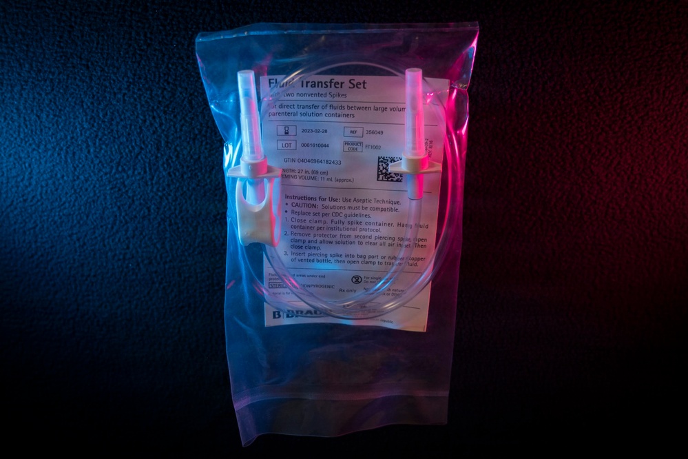 A sealed medical fluid transfer set manufactured by B. Braun is photographed against a dark background with dramatic blue and red lighting. The package contains tubing with two nonvented spikes designed for direct transfer of fluids between large volume parenteral solution containers. The product is still in its original sterile packaging with visible labeling including lot number, date, and usage instructions.