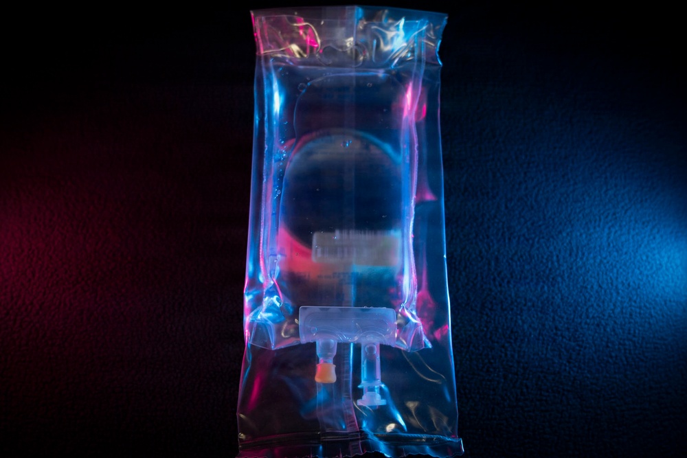 The image shows a medical intravenous (IV) fluid bag photographed under dramatic blue and red/pink lighting against a dark background. The transparent plastic bag contains a clear liquid and features standard IV administration ports at the bottom. The artistic lighting gives the medical equipment a cinematic, stylized appearance.